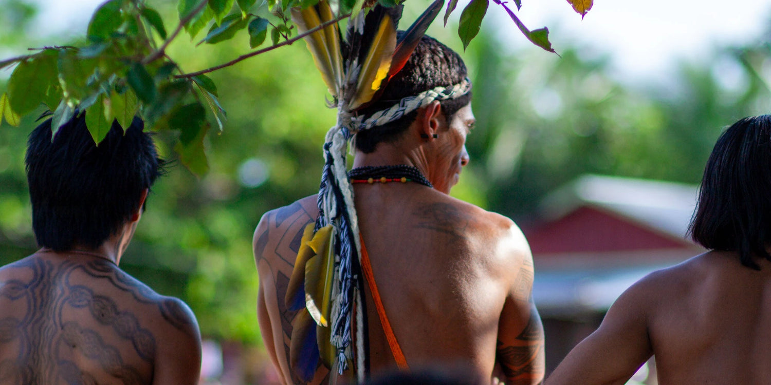 Three tribal men with feathered headdress and temporary festival tattoos on their backs, standing outdoors in the Amazon rainforest