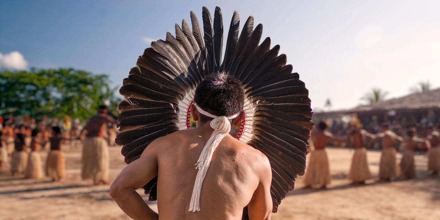 Indigenous man wearing a large feathered headdress, standing in a ceremonial circle with other tribe members in the Amazon